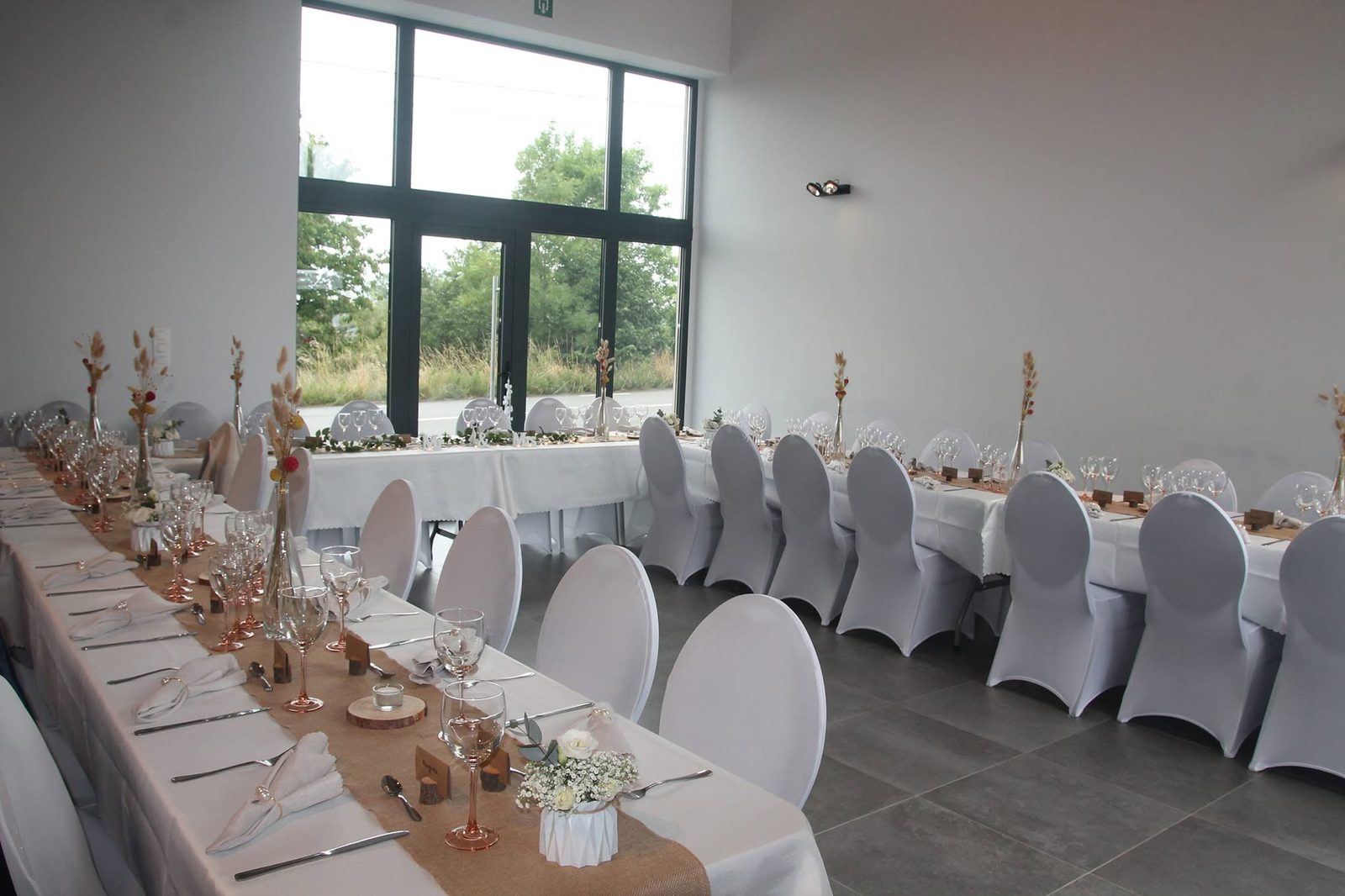 Tables de mariage dressées : housses blanches, chemins de table en jute et baies vitrées sur la campagne
